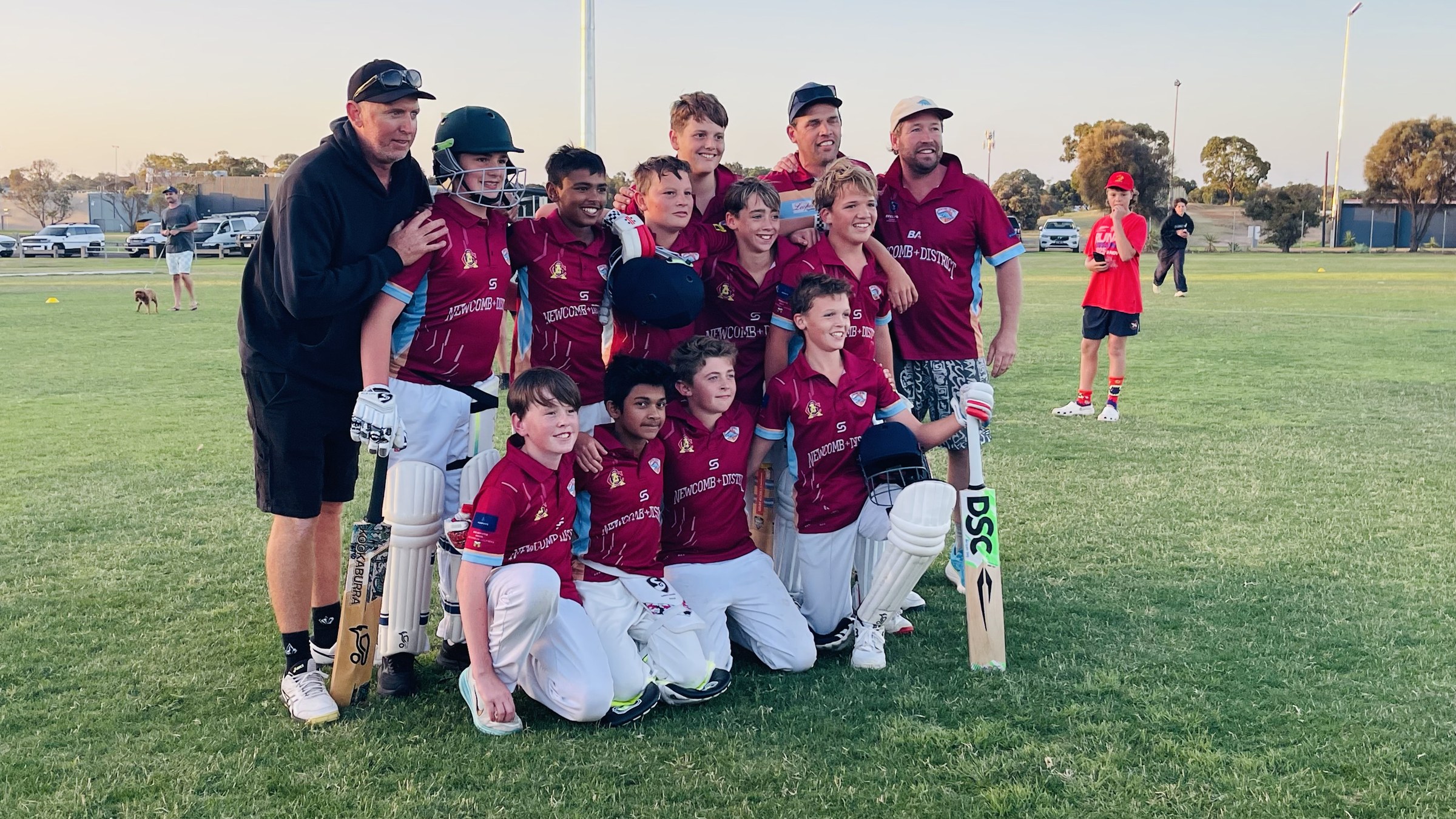 NDCC Under 13 players in maroon shirts and white trousers posing with their coach on the oval.