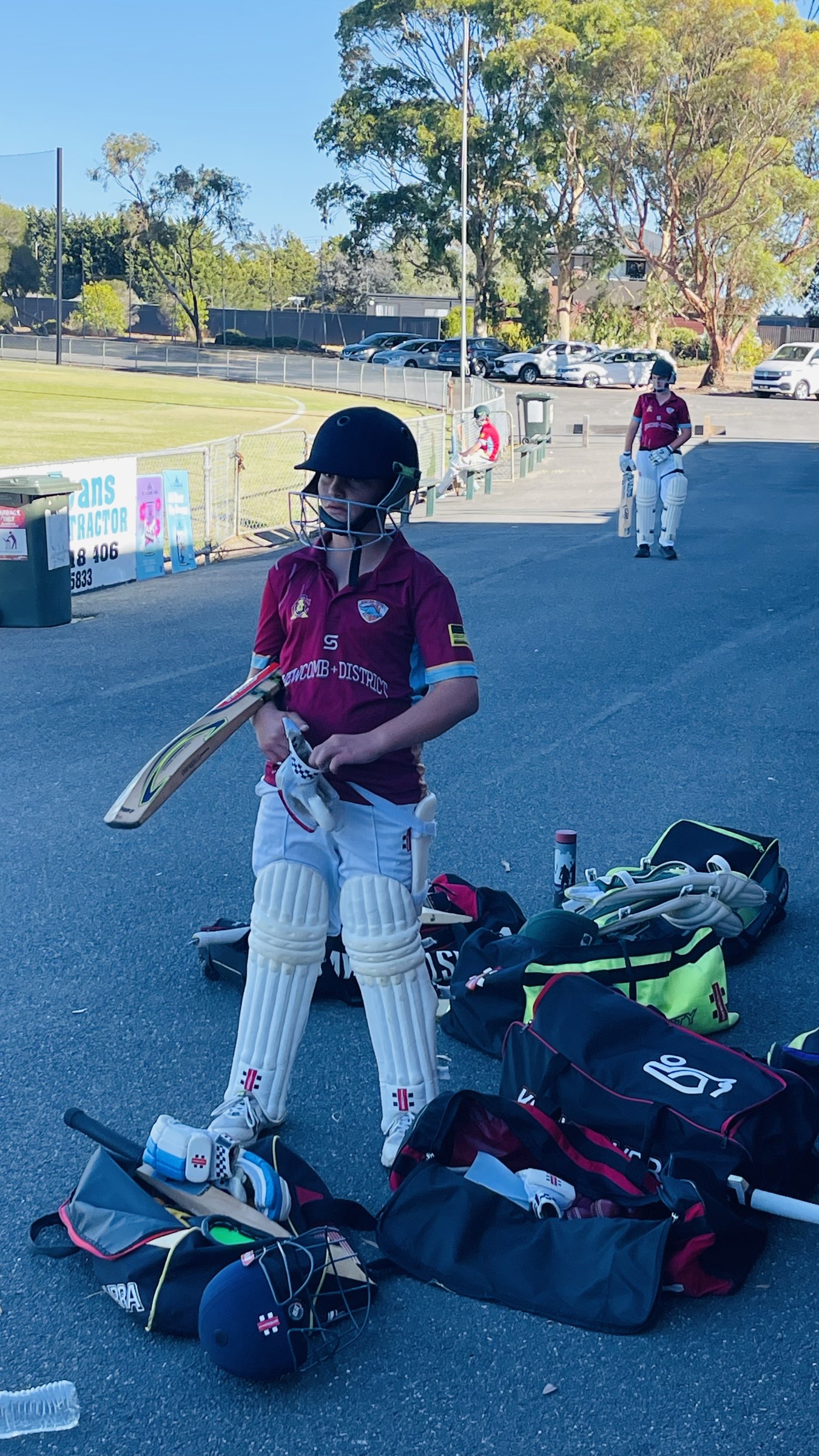 A junior cricketer in maroon kit standing beside cricket bags and equipment.